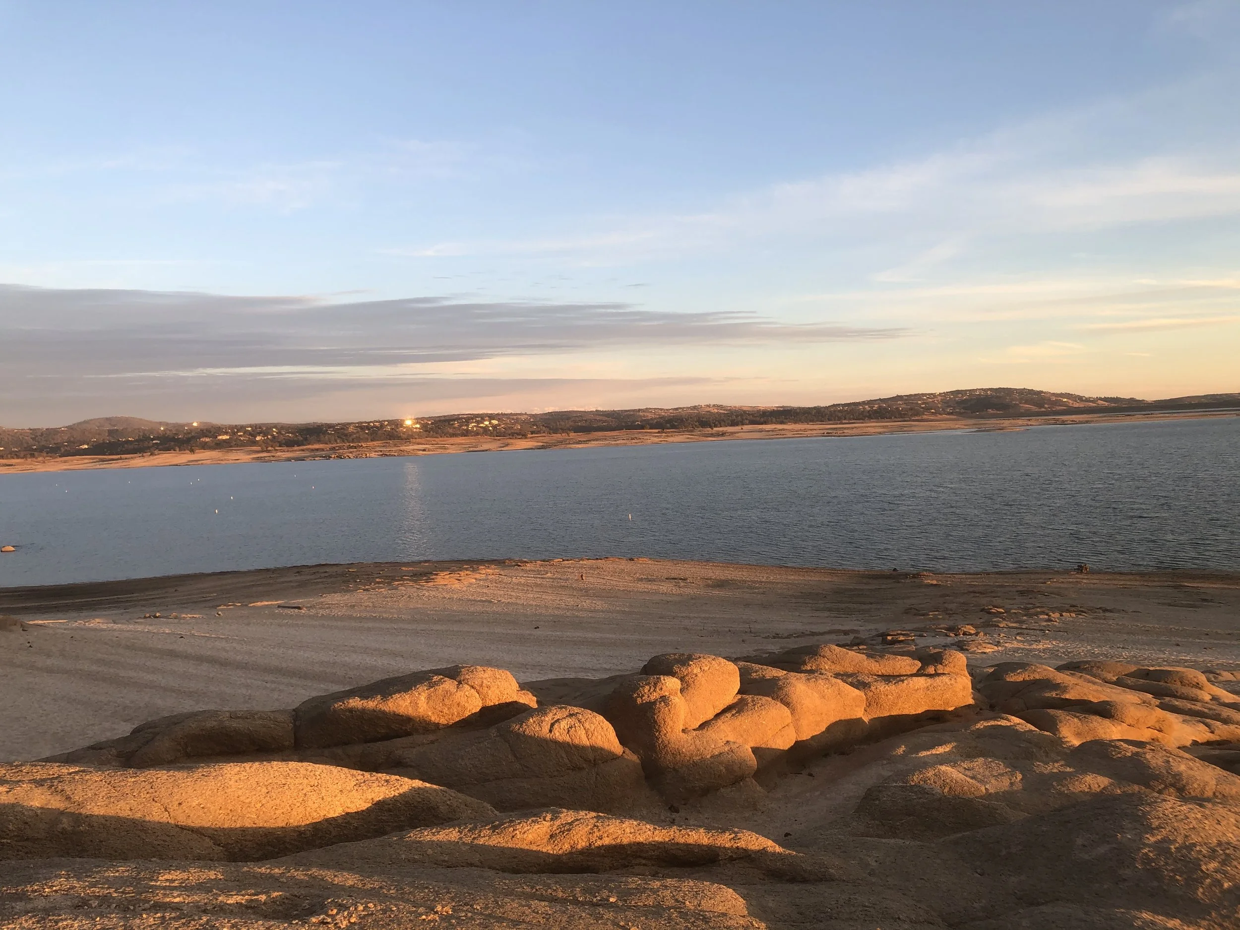 Sunset over a lake with a rocky shoreline in the foreground and hills in the background.
