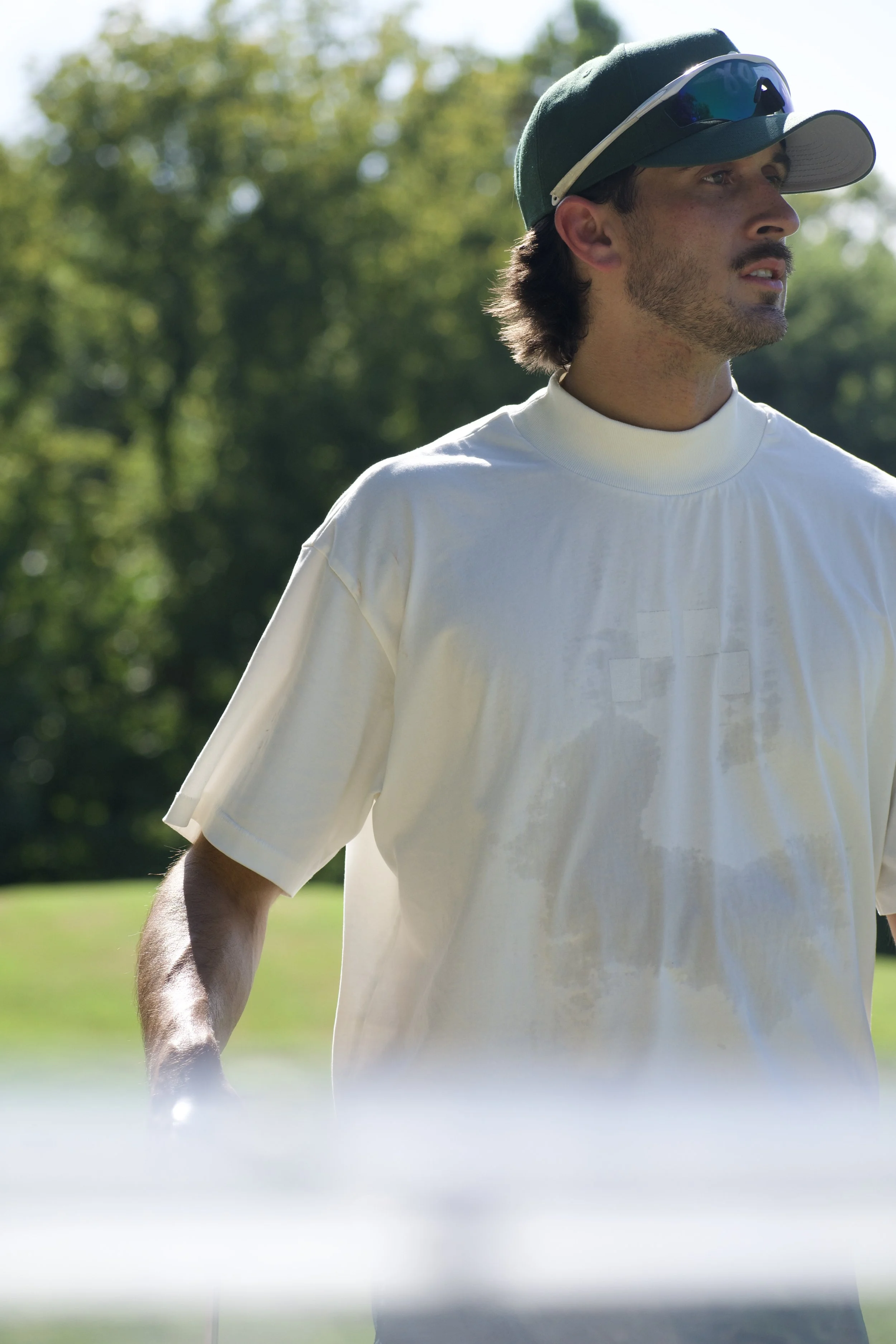 A man dressed as a golfer, wearing a white golf shirt and a dark green cap with sunglasses, standing outdoors on a sunny day with trees in the background.