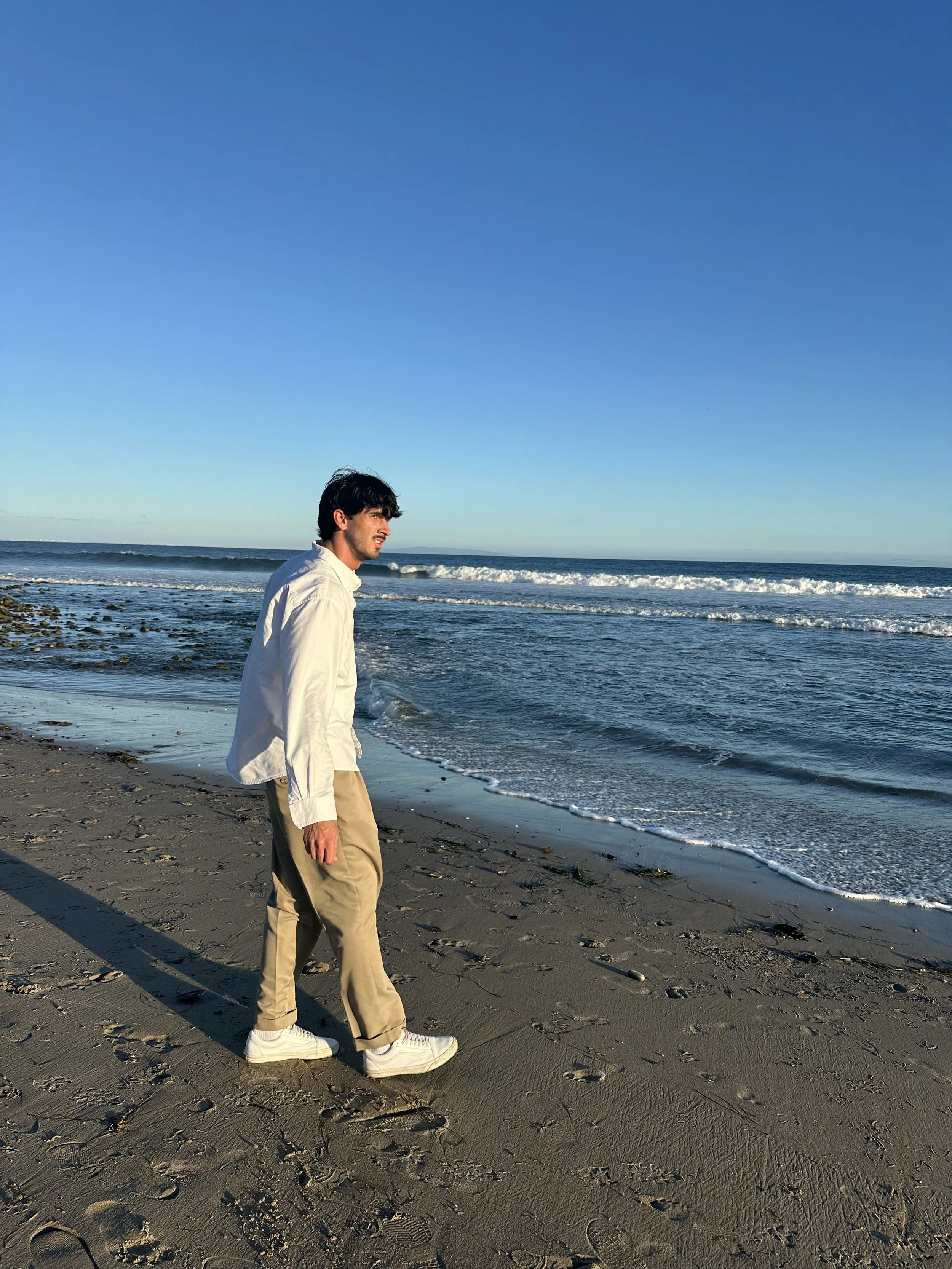 A man walking on the beach near the ocean during sunset or sunrise, wearing a white shirt, beige pants, and white sneakers.