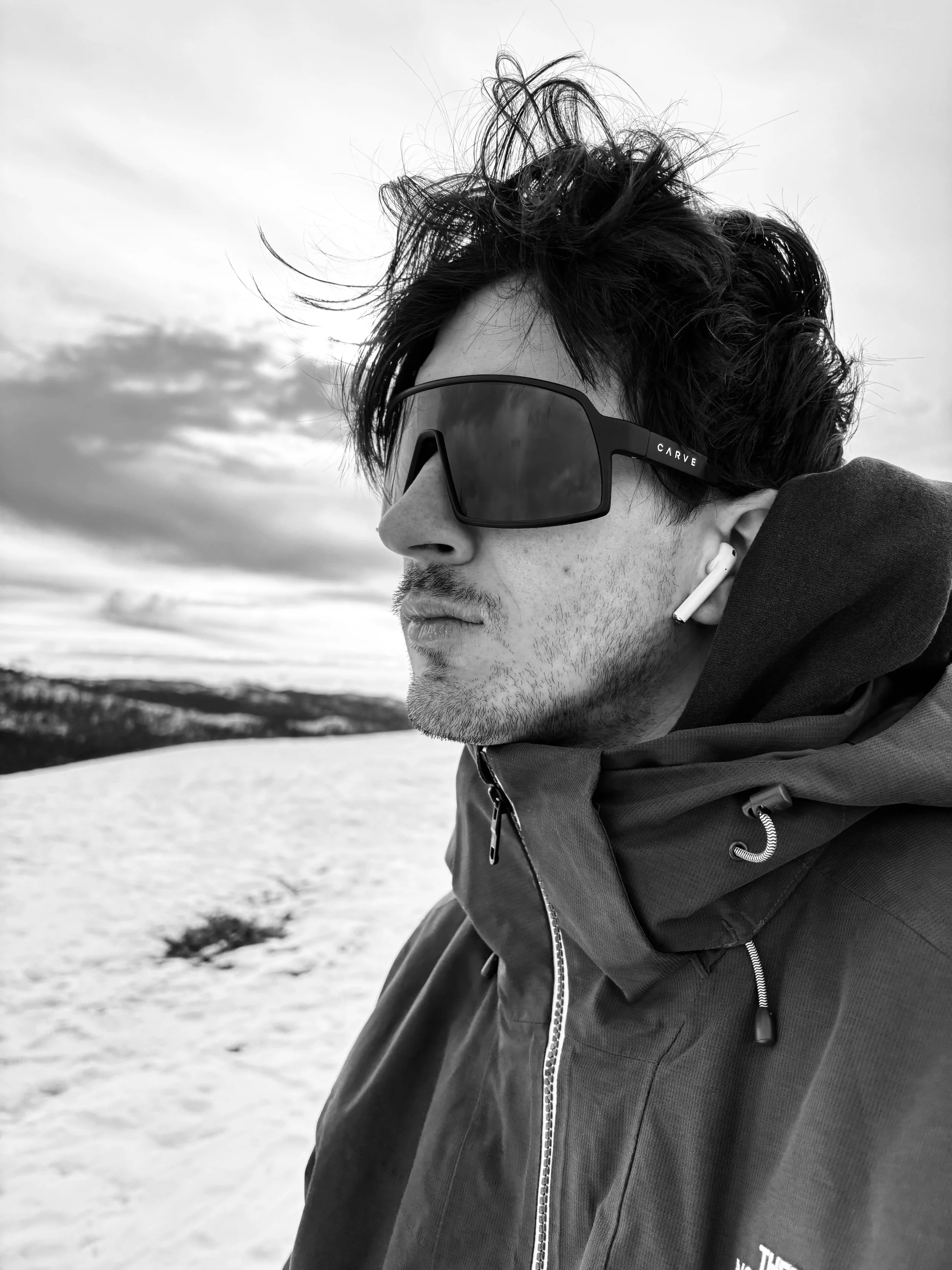 A man outdoors in snowy terrain wearing large sunglasses, a winter jacket with a hood, and wireless earbuds, looking to the side with windswept hair.