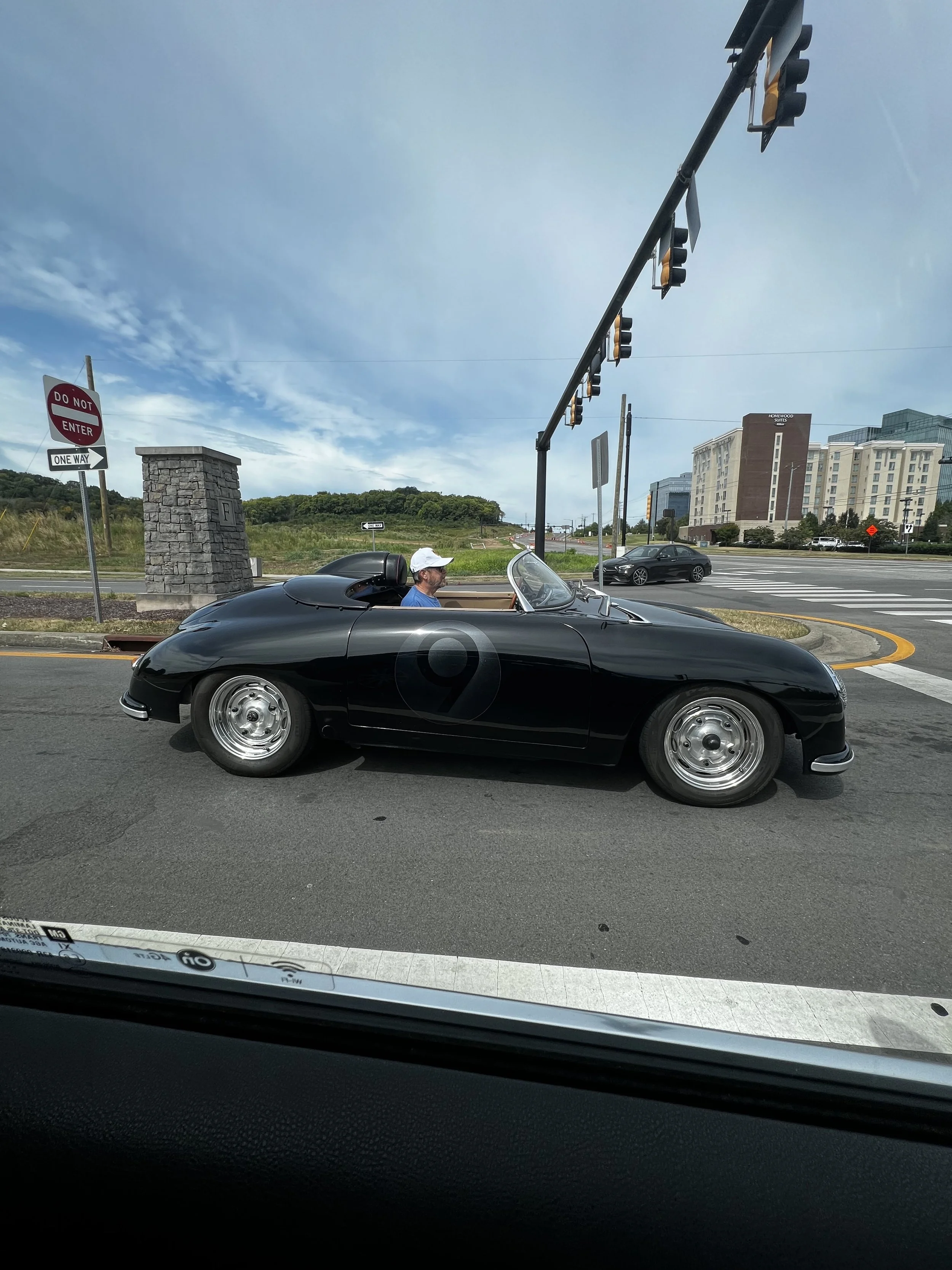 A black vintage convertible car with a man sitting inside, parked at a traffic light on a road in an urban area with buildings and traffic signals in the background.