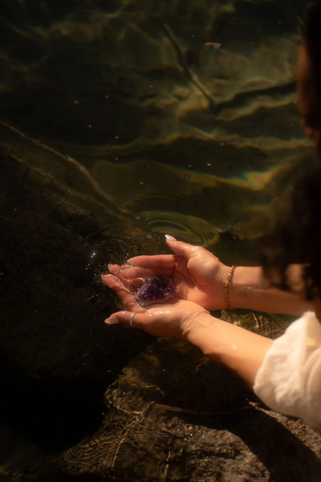 A person’s hands holding a purple gemstone or mineral over a small body of water with rocks surrounding it.