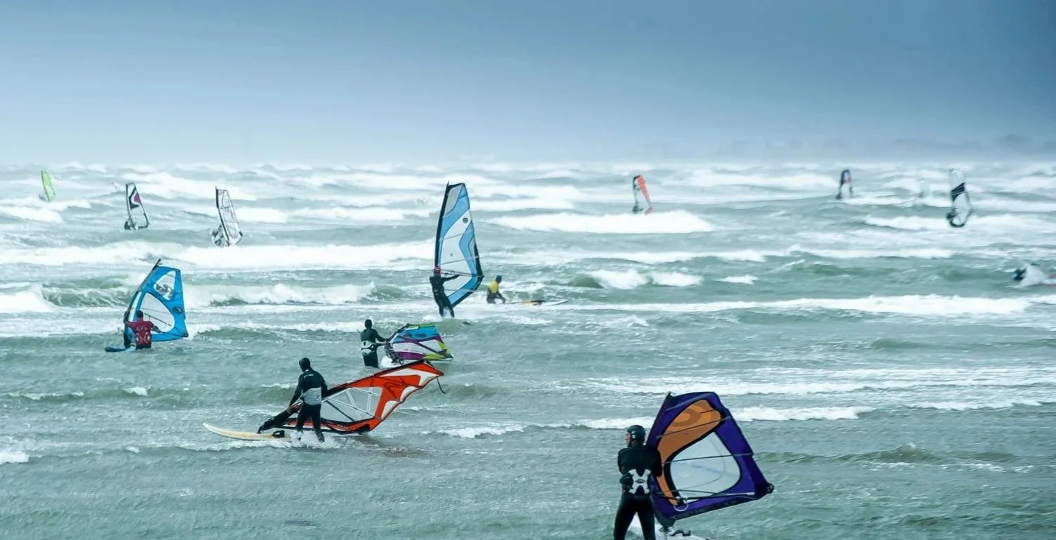 Gruppo di persone che praticano il windsurf in mare, con vele di diversi colori, durante una giornata ventosa.