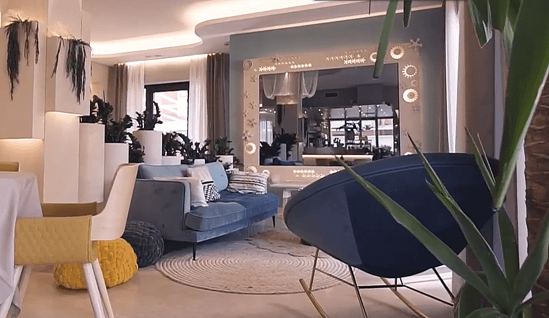 Interior di un soggiorno elegante con divano blu, piante, specchio decorativo e un angolo cucina in background.