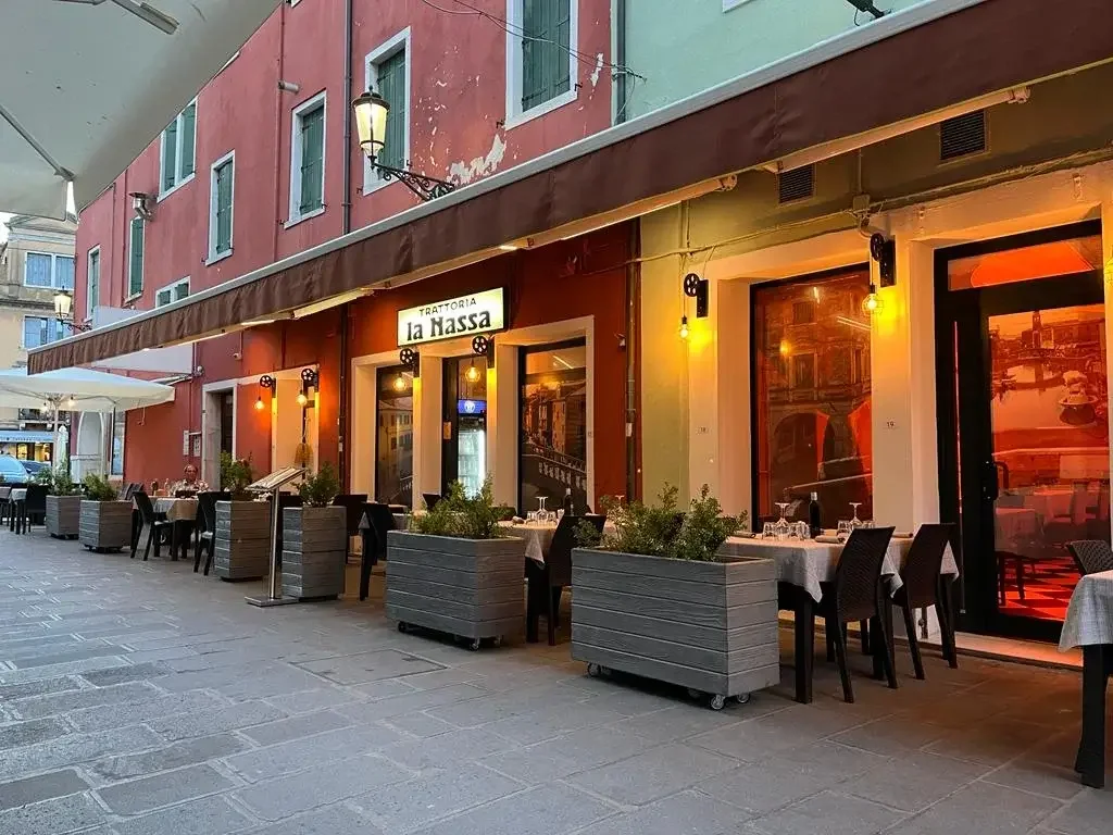Esterno di un ristorante all'aperto in una via urbana con tavoli, sedie e piante. L'illuminazione calda crea un'atmosfera accogliente, con un segno che dice 'Trattoria La Hassa'.