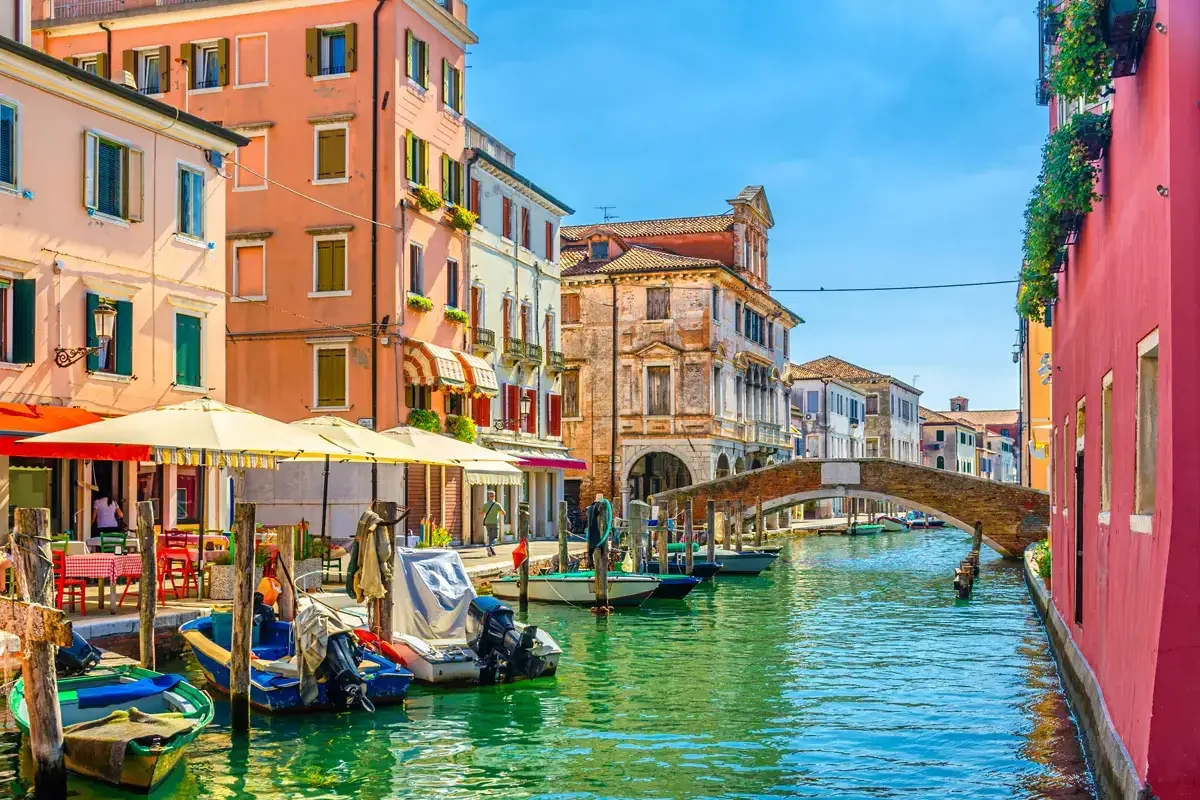 Vista di un canale di Venezia con case colorate che si affacciano sull'acqua, un ponte di pietra, barche ormeggiate e un cielo azzurro.