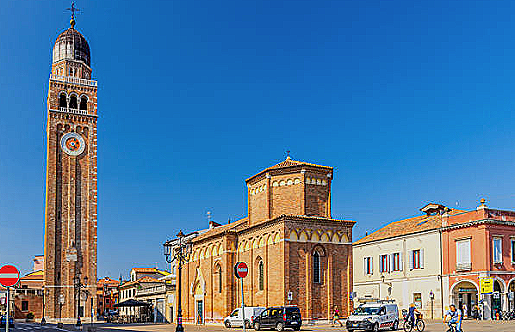 Vista di un campanile e una chiesa nel centro di una città, con edifici dai colori chiari e traffico di auto e biciclette