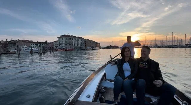Due persone sedute su una barca al tramonto in un porto con edifici e altre imbarcazioni sullo sfondo.