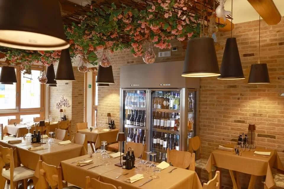 Interno di un ristorante arredato con tavoli con tovaglie beige, sedie di legno, illuminazione soffusa con lampade nere, pareti in mattoni e decorazioni floreali sospese al soffitto.