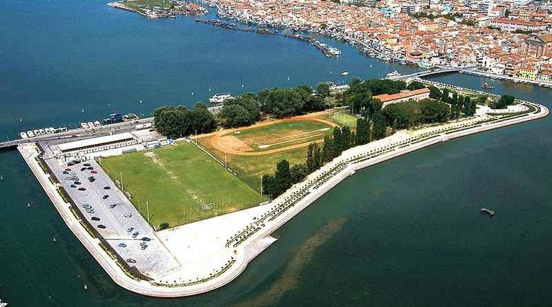 Veduta aerea di un'area portuale con campi sportivi, una strada con auto e un ponte che attraversa il mare, con una città sullo sfondo.