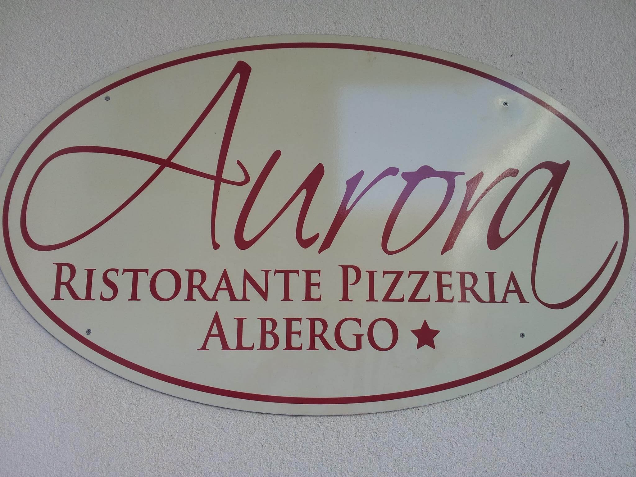 Segno o targhetta con scritto 'Aurora Ristorante Pizzeria Albergo' in caratteri rossi su uno sfondo bianco e un'ellisse rossa che circonda la scritta.