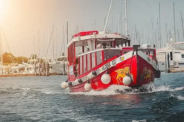 Immagine di una nave rossa che percorre un porto, con altre barche ormeggiate sullo sfondo.