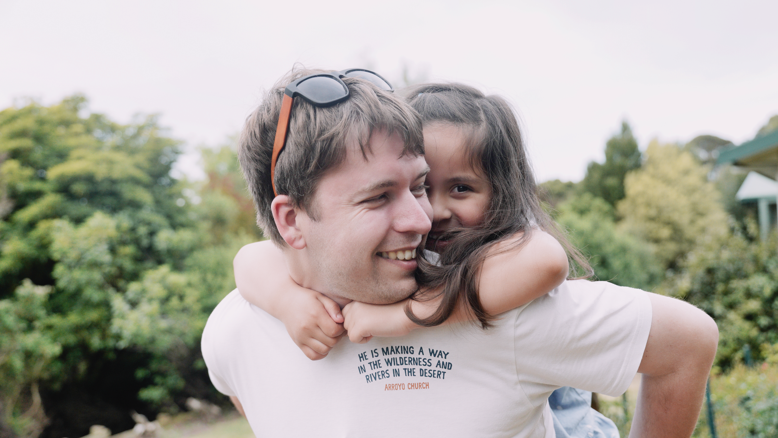 A young man with sunglasses on his head giving a piggyback ride to a girl with long dark hair, both smiling outdoors with green trees in the background.