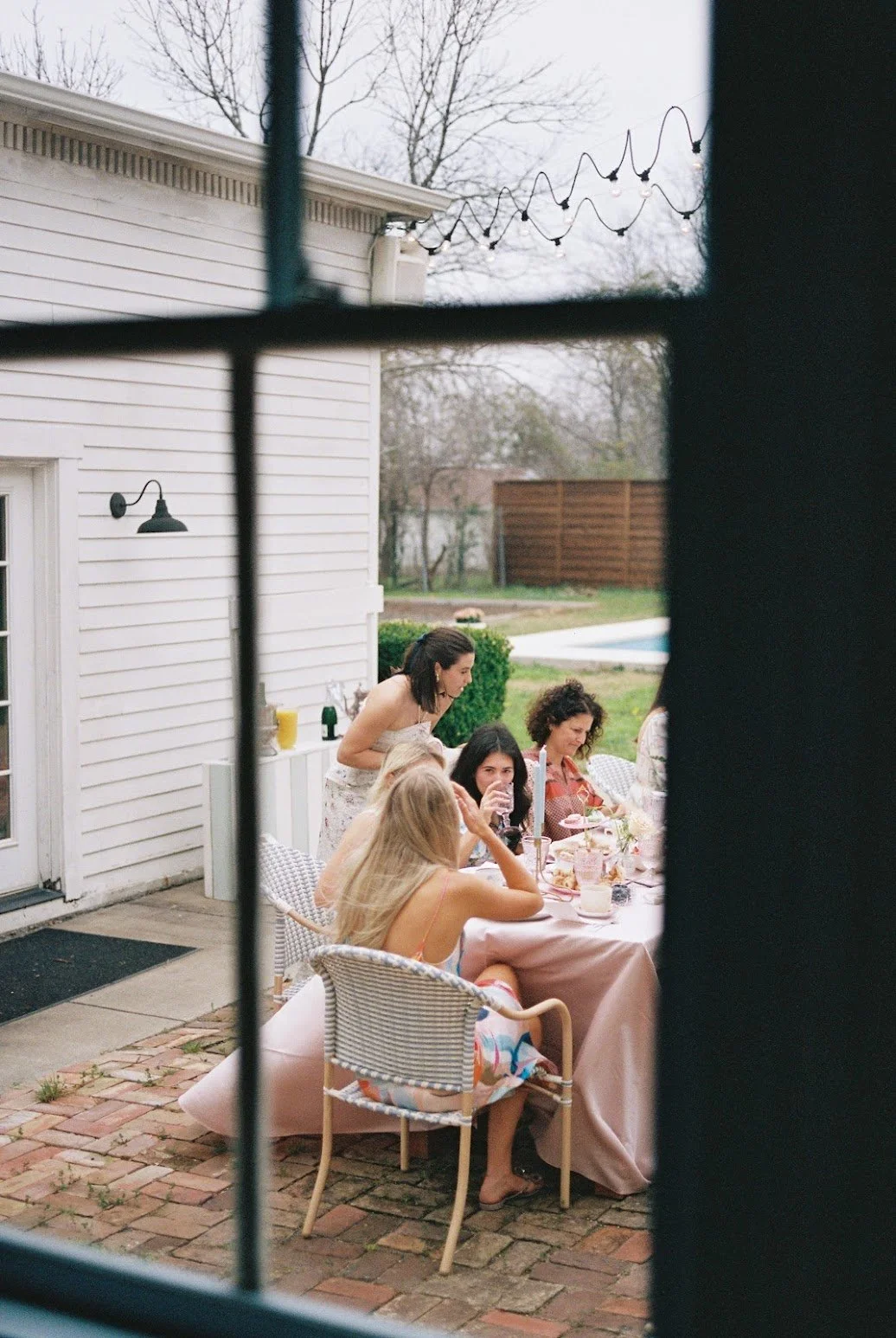 A group of women gathered around a table outside, enjoying a social occasion, seen through a window.