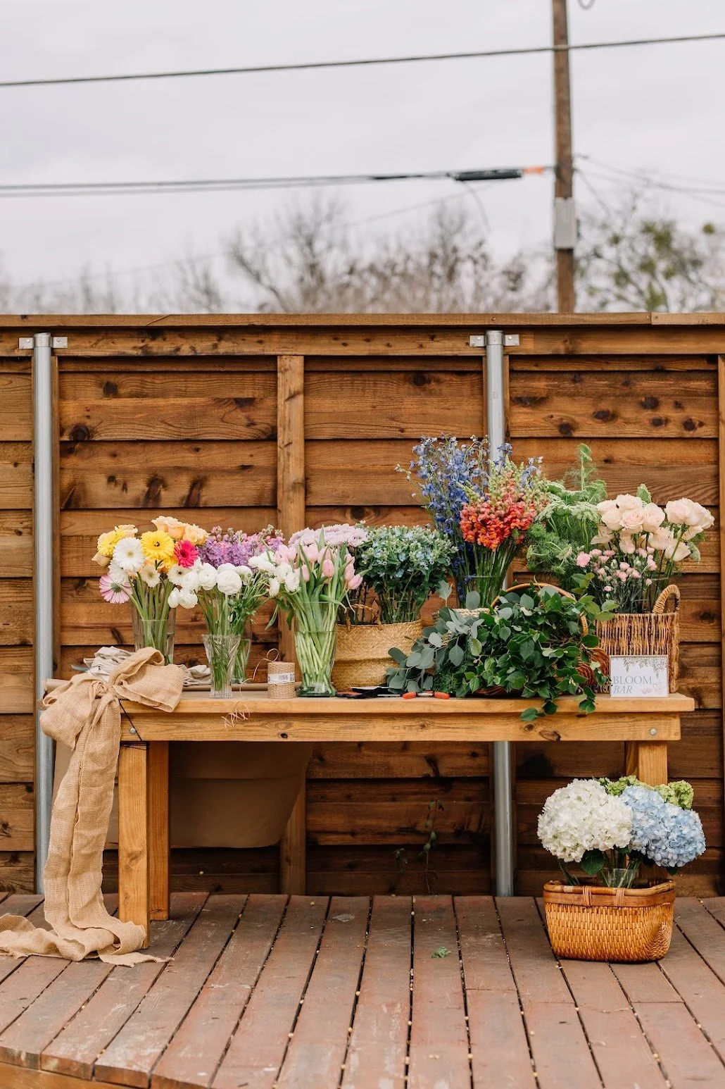 Flowers arranged on a wooden table in front of a wooden fence, with a small sign reading 'Bloom Bar' and a basket of hydrangeas on the ground.