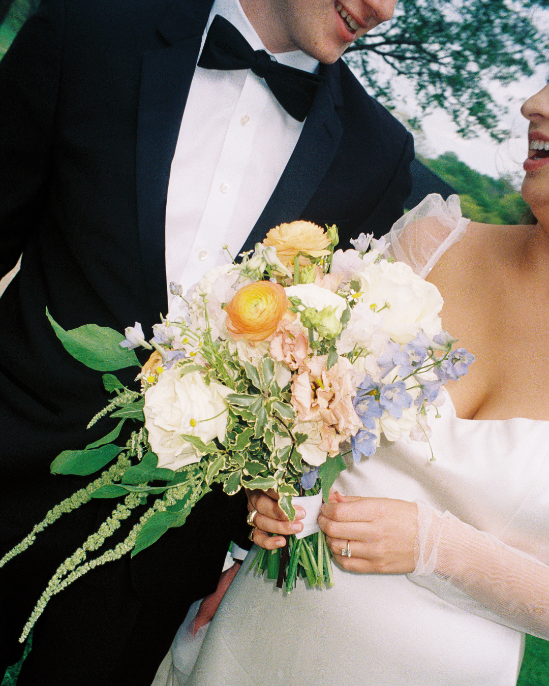 Close-up of a newlywed couple holding a bouquet of flowers. The groom is in a tuxedo with a black bowtie, and the bride is in an off-the-shoulder wedding dress with sheer sleeves.