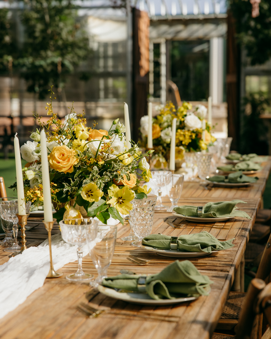 Decorated outdoor dining table with floral centerpieces, candles, glassware, and green napkins.
