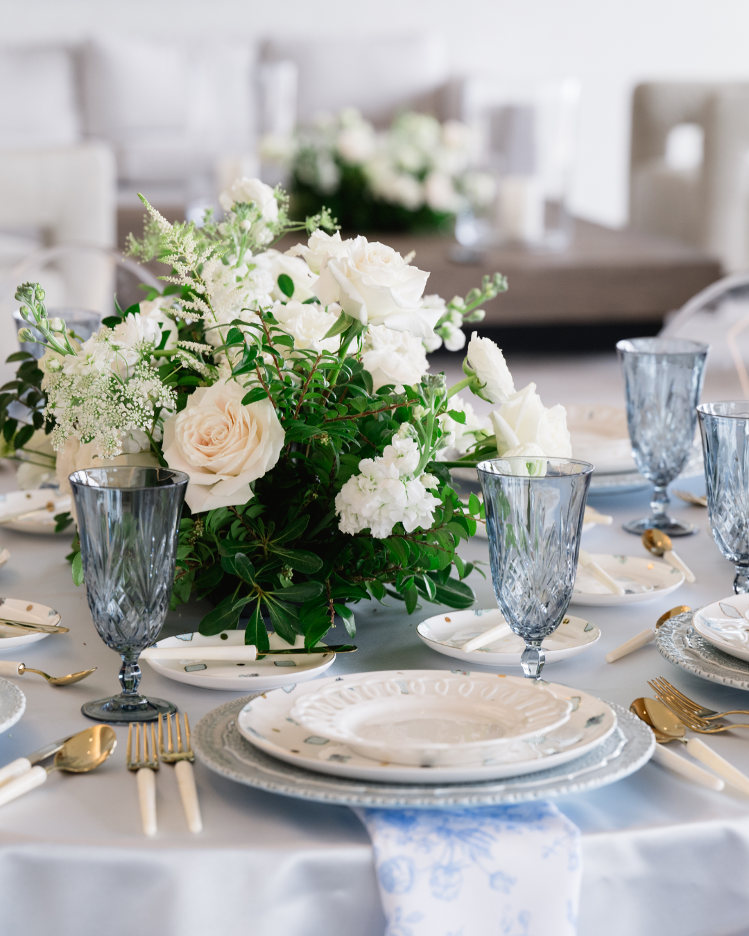 Elegant dining table with a white floral centerpiece, blue glassware, and white china plates with gold utensils, set for a formal event.
