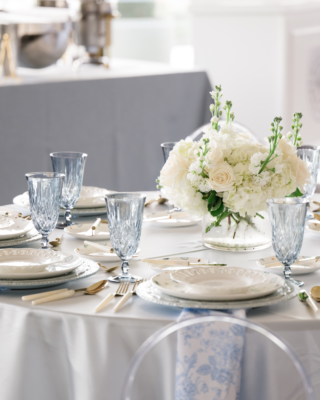 Elegant table setting with white and gold dishware, crystal glasses, and a floral centerpiece in a glass vase with white roses and greenery.