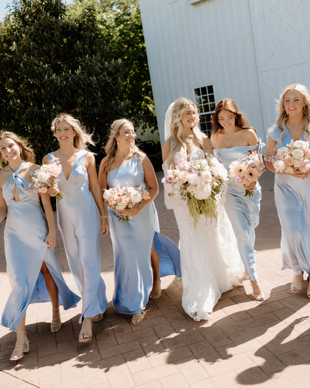 A bride and her five bridesmaids walking outdoors on a sunny day, holding bouquets of flowers, with trees and a white building in the background.