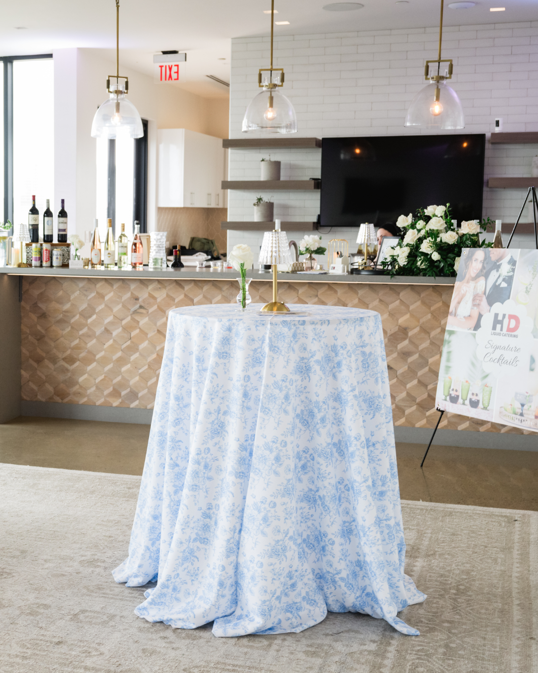Interior of a modern event space with a round table covered in a blue floral tablecloth, featuring a small lamp, and a backdrop of a bar with various bottles, flowers, and signage for signature cocktails.