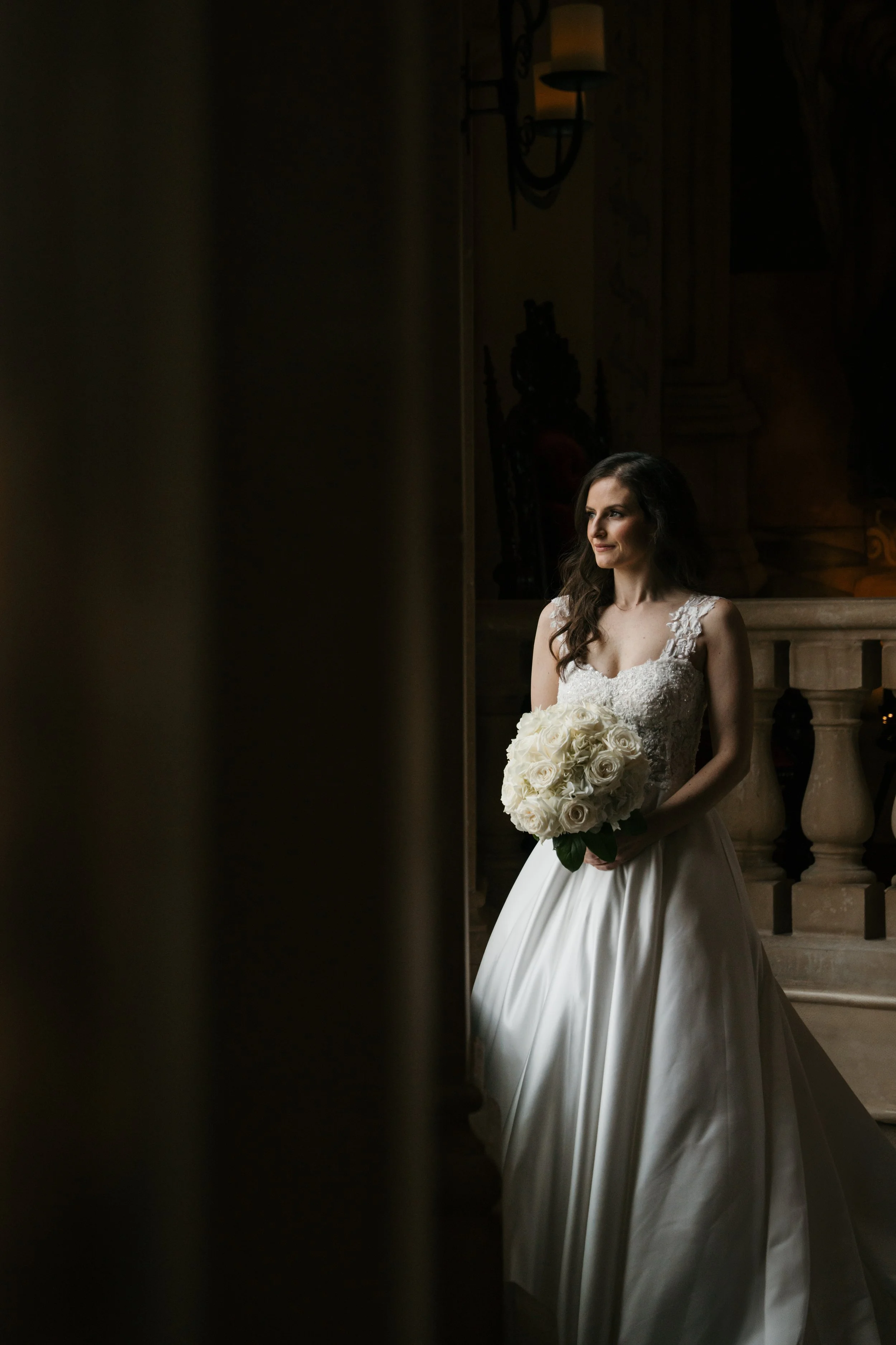 A bride in a wedding dress holding a bouquet of white roses, standing indoors near a staircase with ornate railing, softly lit with warm lighting.