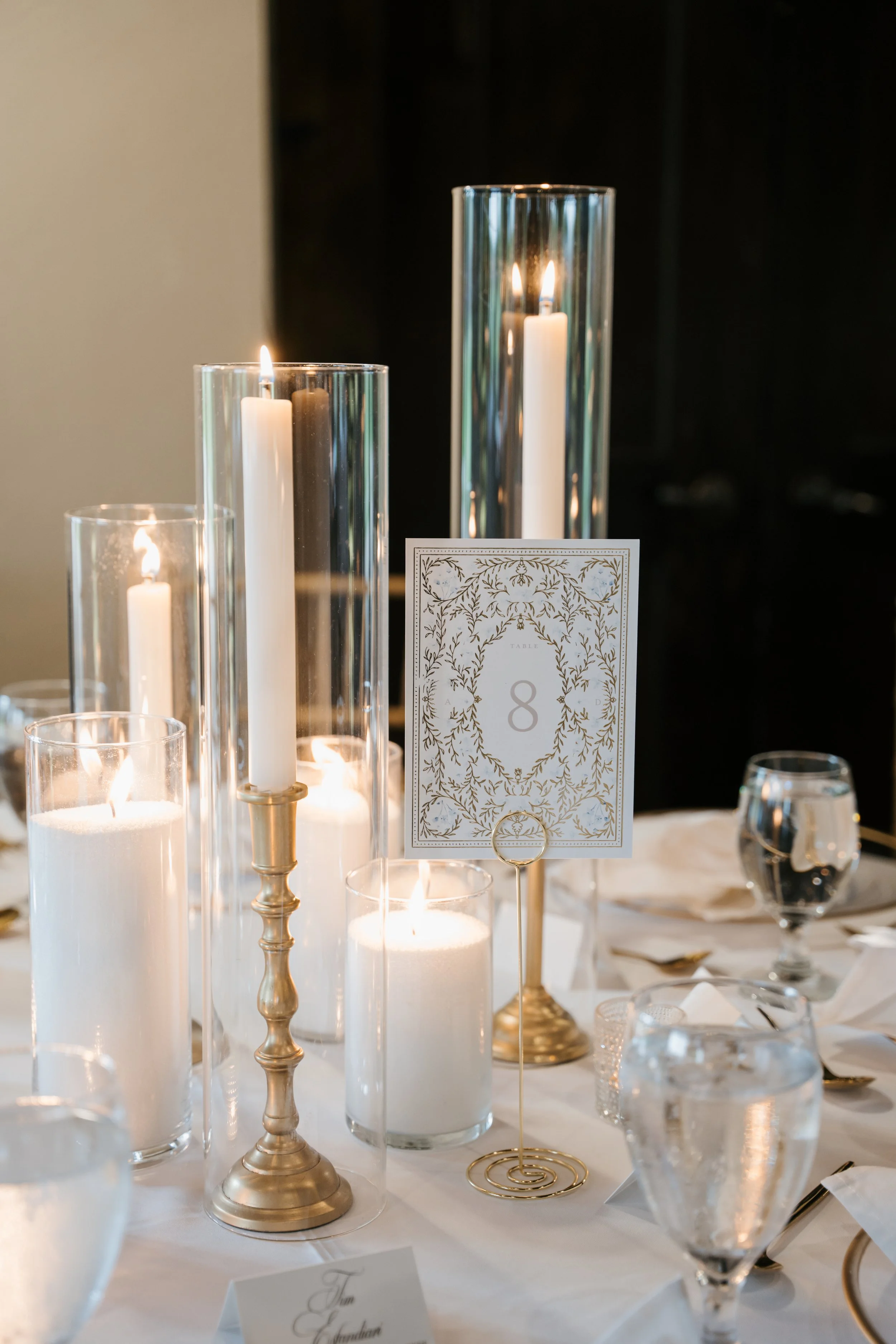 Elegant table centerpiece with lit white candles in tall glass holders, gold accents, table number 8 card, and water glasses on a white tablecloth.