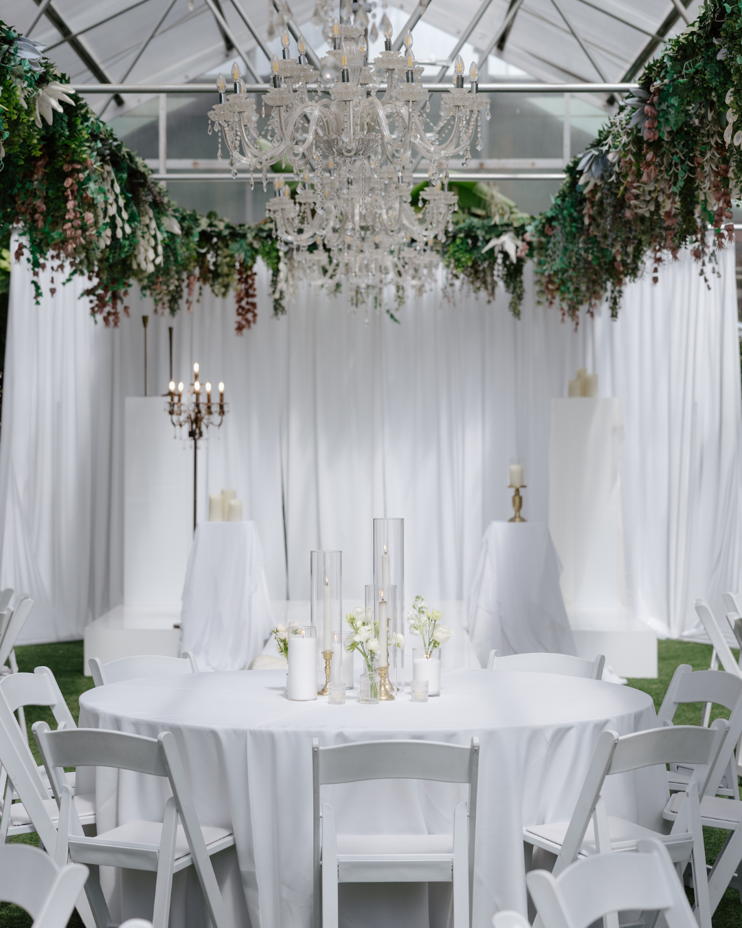 Elegant event space with a round table covered in white cloth, surrounded by white chairs, adorned with white flowers in vases, multiple tall glass candle holders with candles, and surrounded by draped white curtains, hanging chandeliers, and floral
