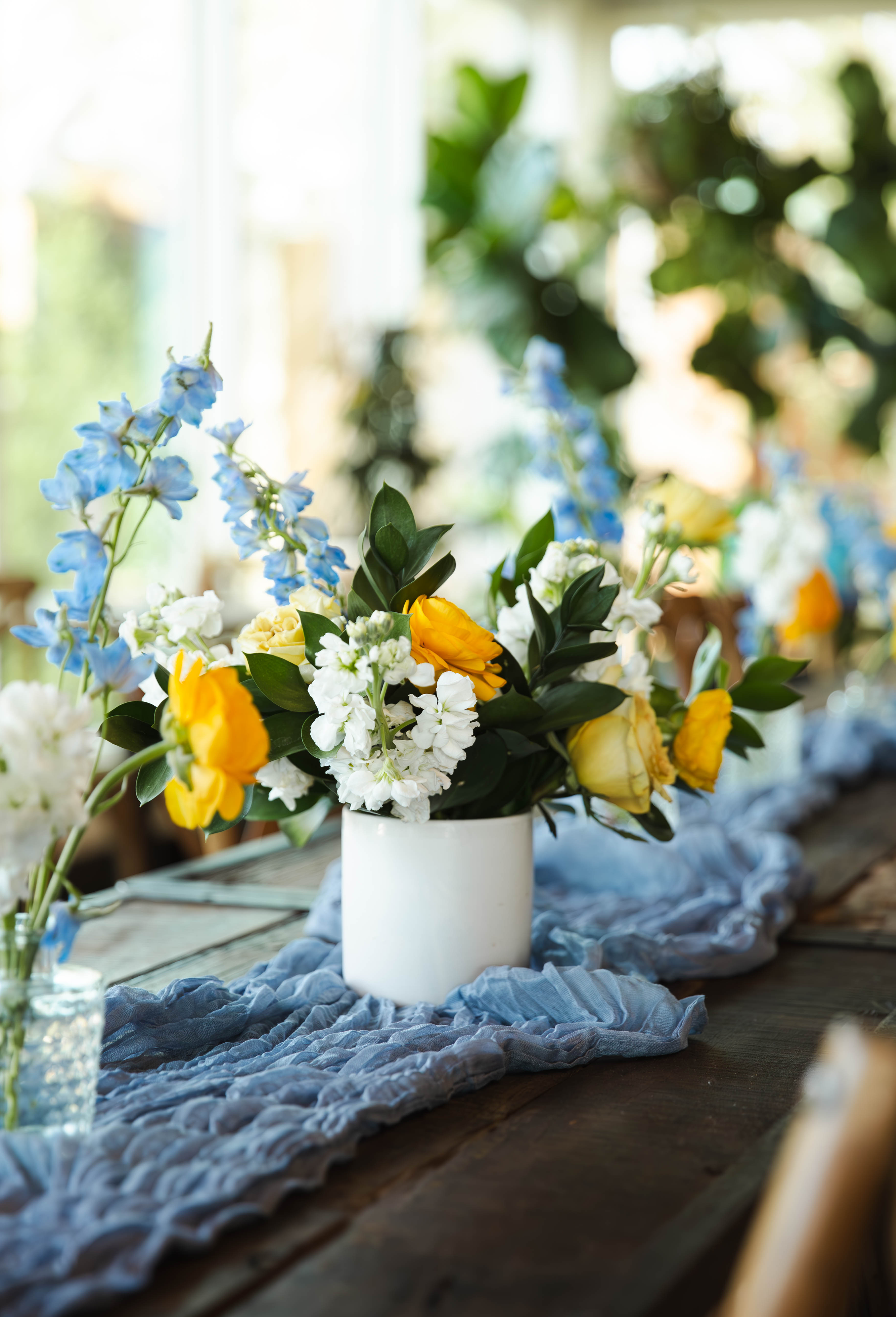 A white vase with a colorful flower arrangement, including yellow roses, white flowers, and blue delphiniums, on a table with a blue cloth runner.