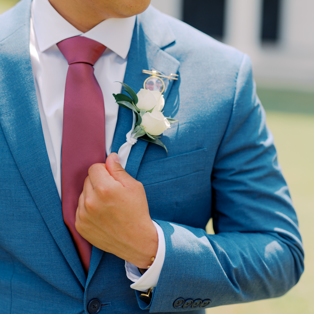 Close-up of a man in a blue suit adjusting a pink tie, with a white boutonniere and a small photo badge pinned on his lapel.