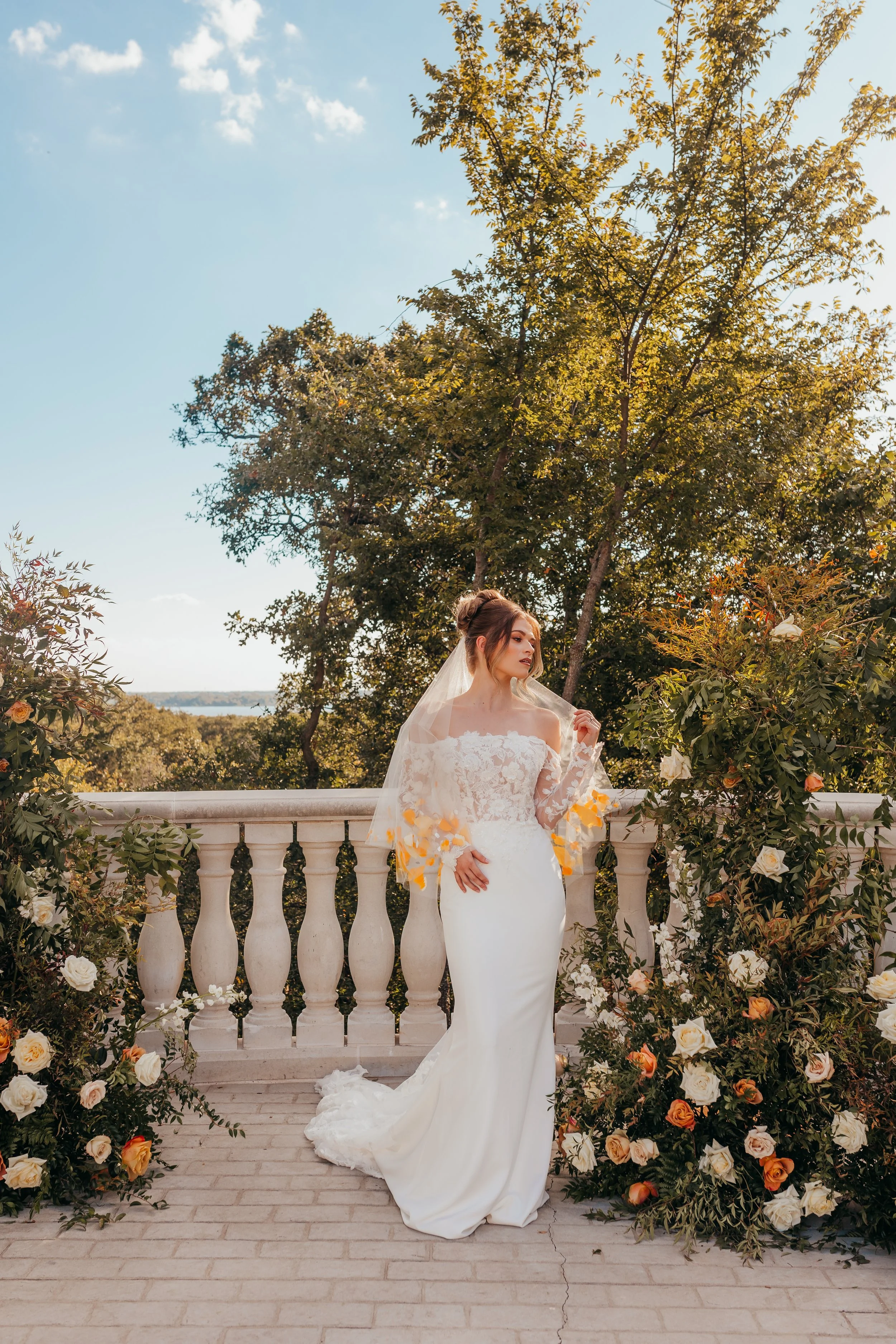 A bride in a white wedding gown with lace details on the off-shoulder top and long sleeves, standing on a balcony with a white balustrade, surrounded by flower arrangements, outdoors with green trees and a blue sky in the background.