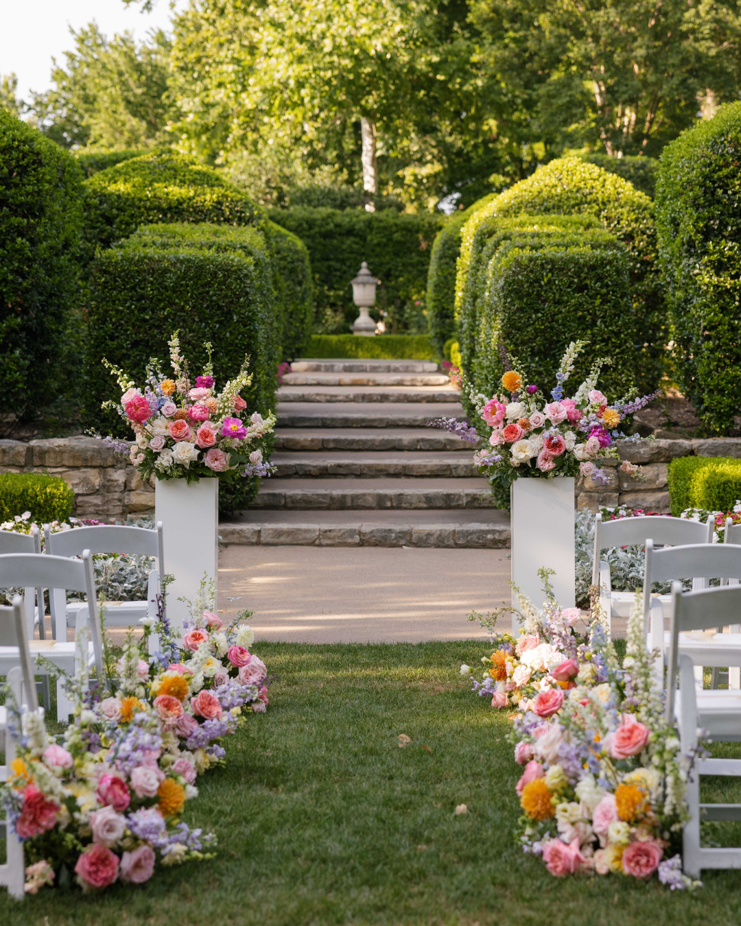 Outdoor wedding ceremony setup with white chairs, floral arrangements, and stone steps leading to a garden area.