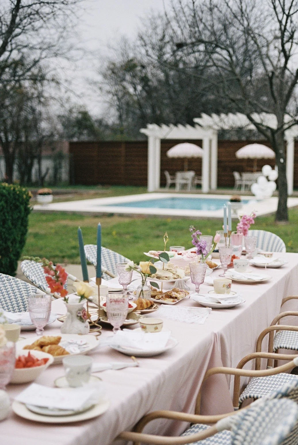 An outdoor garden party setup with a long table decorated with pink linens, flowers, candles, and tableware, set near a swimming pool with patio furniture and umbrellas in the background.