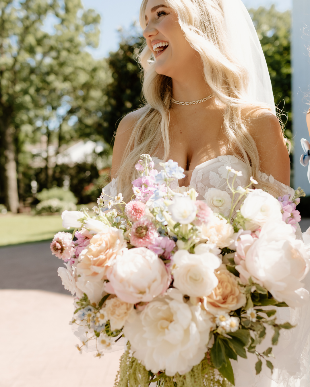 A smiling woman with long blonde hair in a wedding dress holding a large bouquet of pastel-colored flowers outdoors.