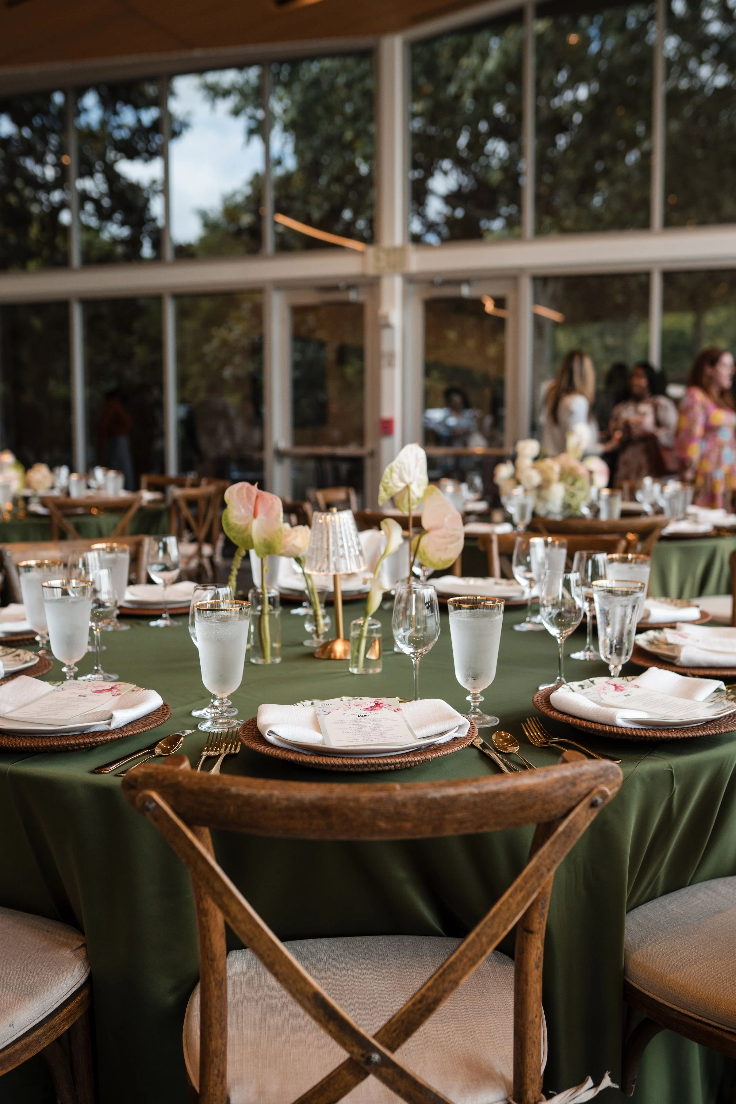Elegant event table setting with green tablecloth, floral centerpieces, wine glasses, water glasses, plates with napkins, and cutlery, in a glass-enclosed venue with guests in the background.