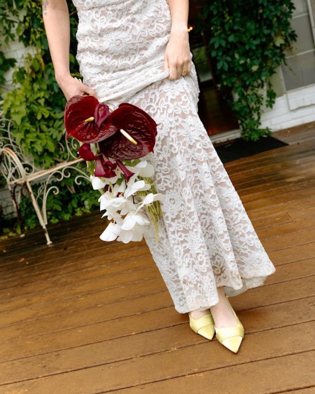 A person wearing a white lace dress is holding a bouquet of red and white flowers. They are standing on a wooden deck outside, with green plants and a white brick wall in the background.