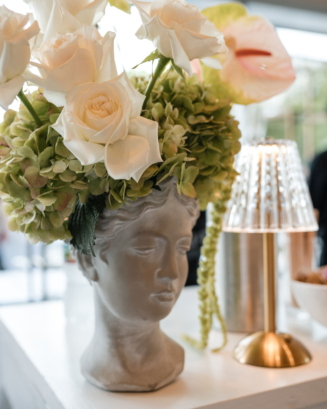 A decorative bust of a woman with closed eyes, used as a vase for a floral arrangement of white roses, hydrangeas, and other flowers. A table lamp with a gold base and clear shade is visible in the background.
