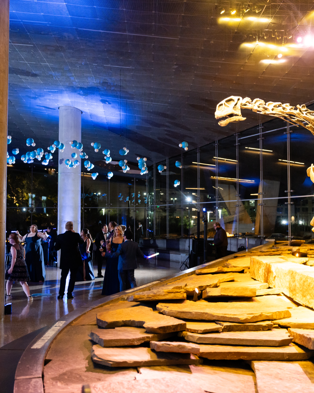 A group of people in formal attire dancing and socializing inside a modern building with a high ceiling, large glass windows, blue and white balloons suspended from the ceiling, and decorative stone elements in the foreground.