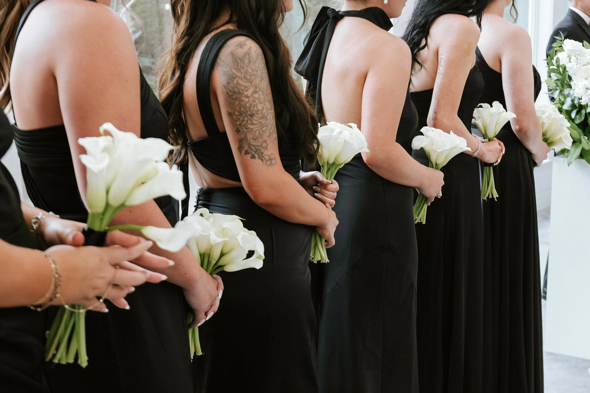 Line of women in black dresses holding white calla lily bouquets at a wedding or formal event.