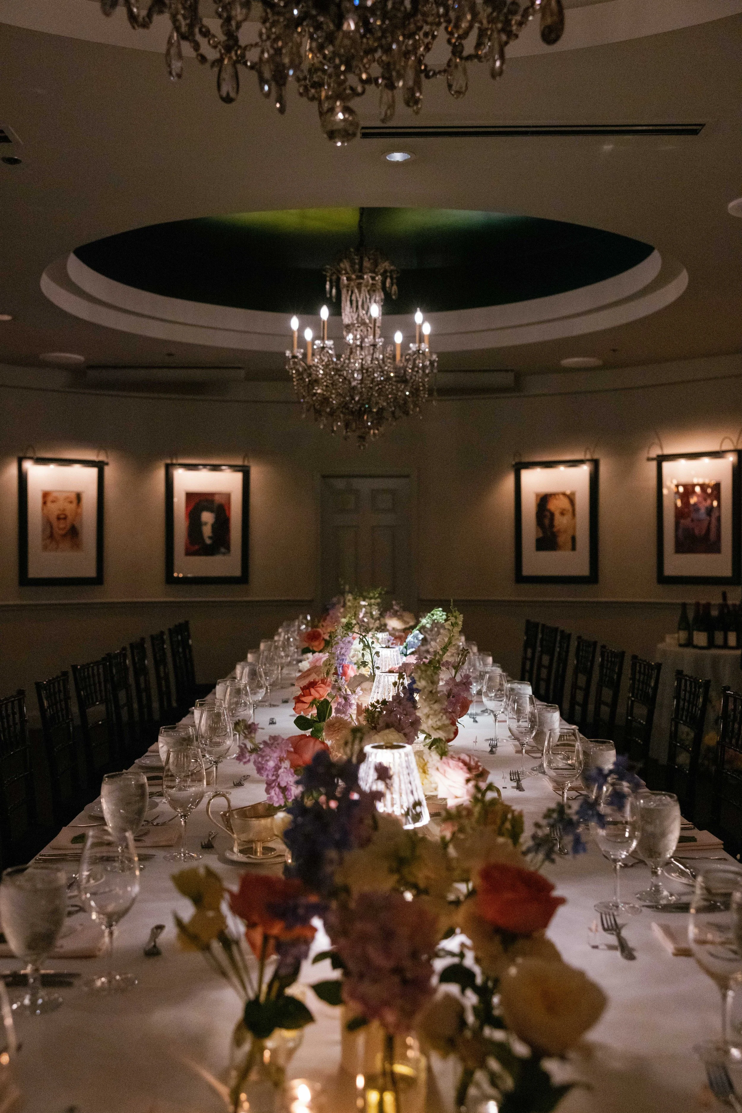 Elegant banquet table decorated with flowers, lit by small lamps, in a dimly lit room with framed artwork and chandeliers.