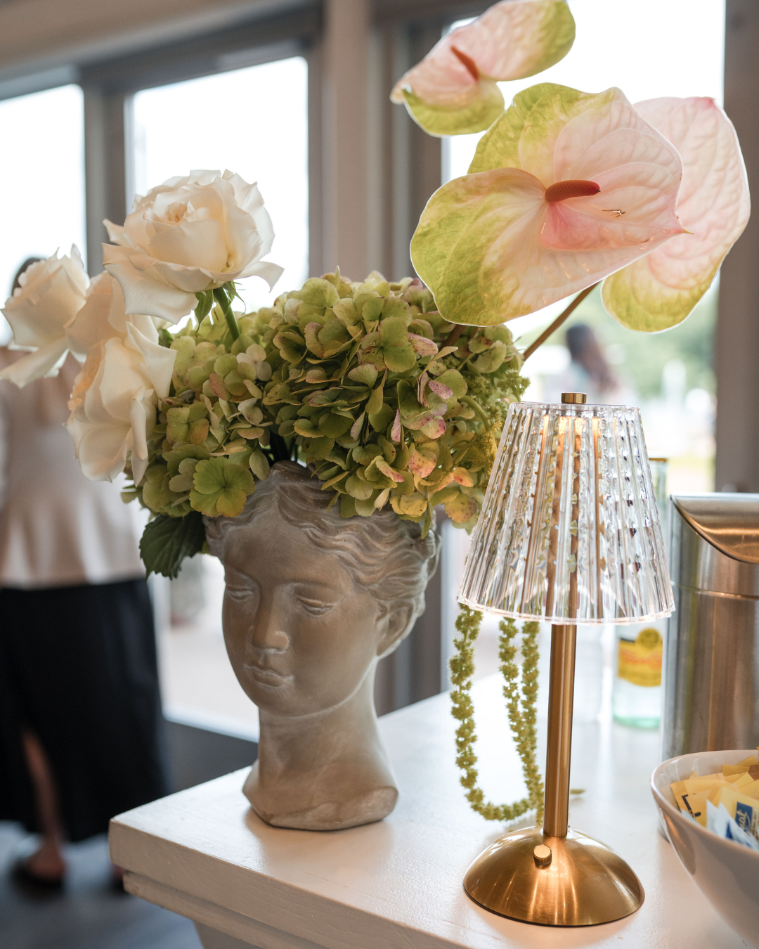 A decorative flower arrangement in a bust-shaped vase featuring white roses, green hydrangeas, and pink-tinged anthuriums, with a brass table lamp beside it.