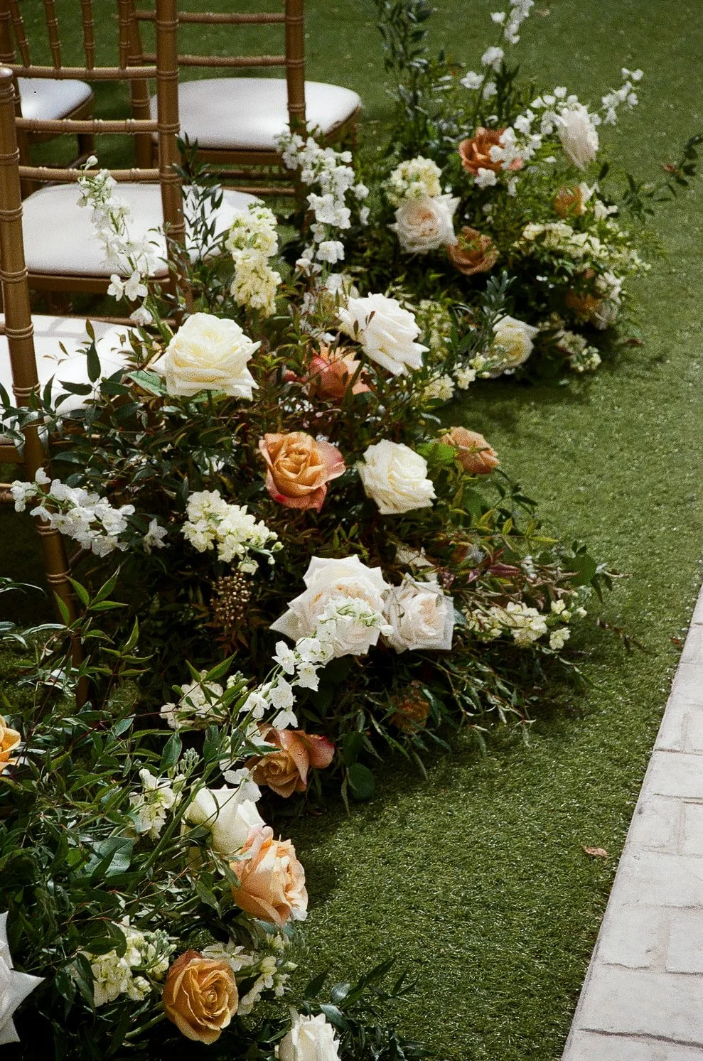 Wedding aisle decorated with lush floral arrangements featuring white and peach roses, white hydrangeas, and greenery, with gold chairs lining the aisle on a green carpet.