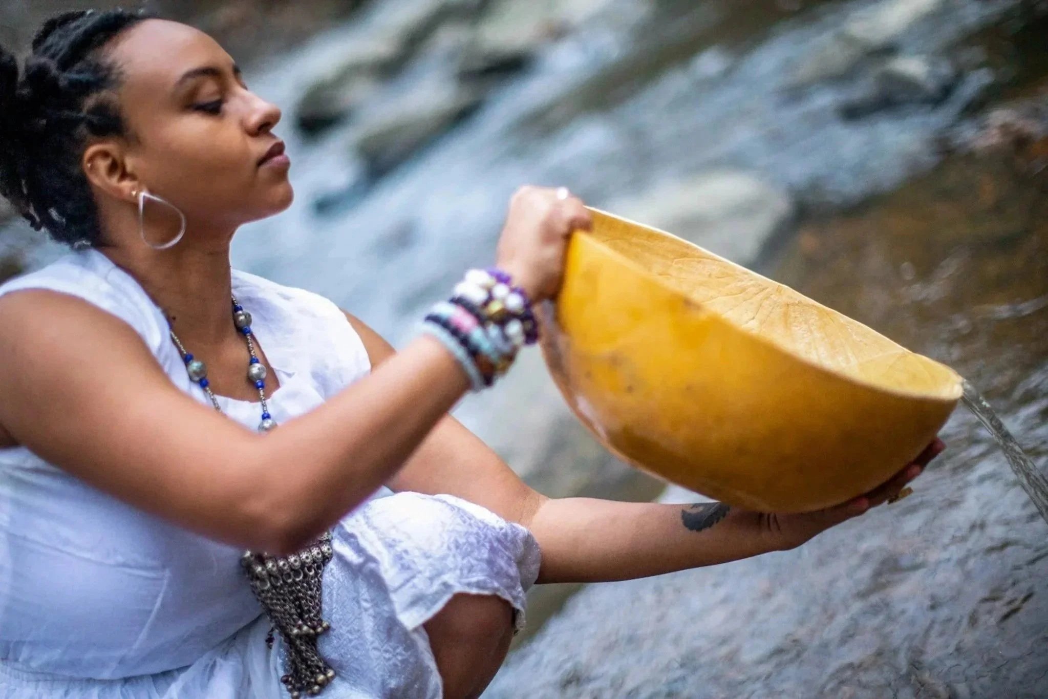 A woman in white clothing and jewelry is sitting by a creek, holding a large, yellow wooden bowl, with water flowing into it.