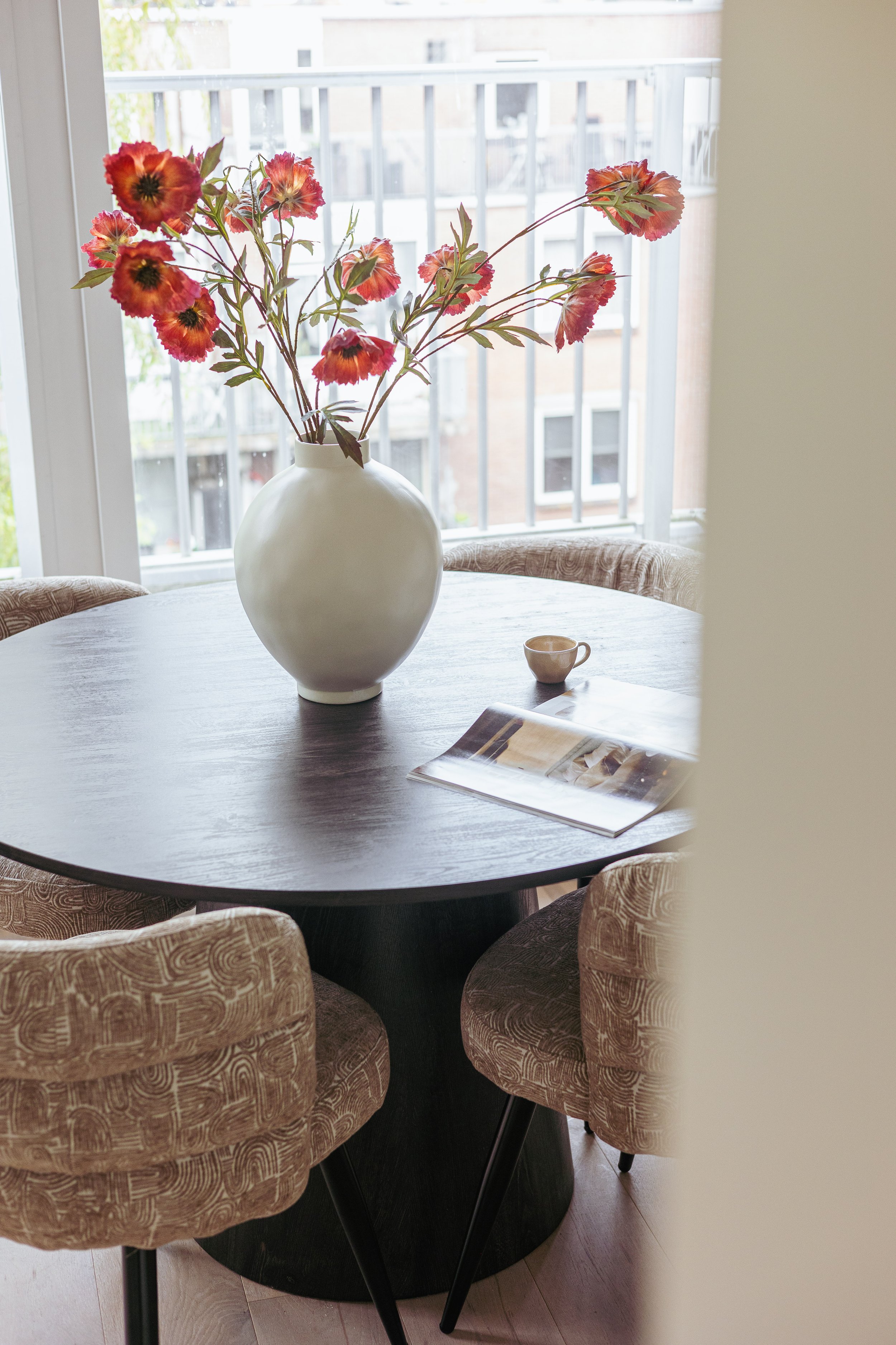 Een ronde houten tafel met een grote witte vaas met rode en roze bloemen, een kleine kopje en een magazine, omringd door vier beige beklede stoelen, bij een raam dat uitkijkt op een buitenruimte.