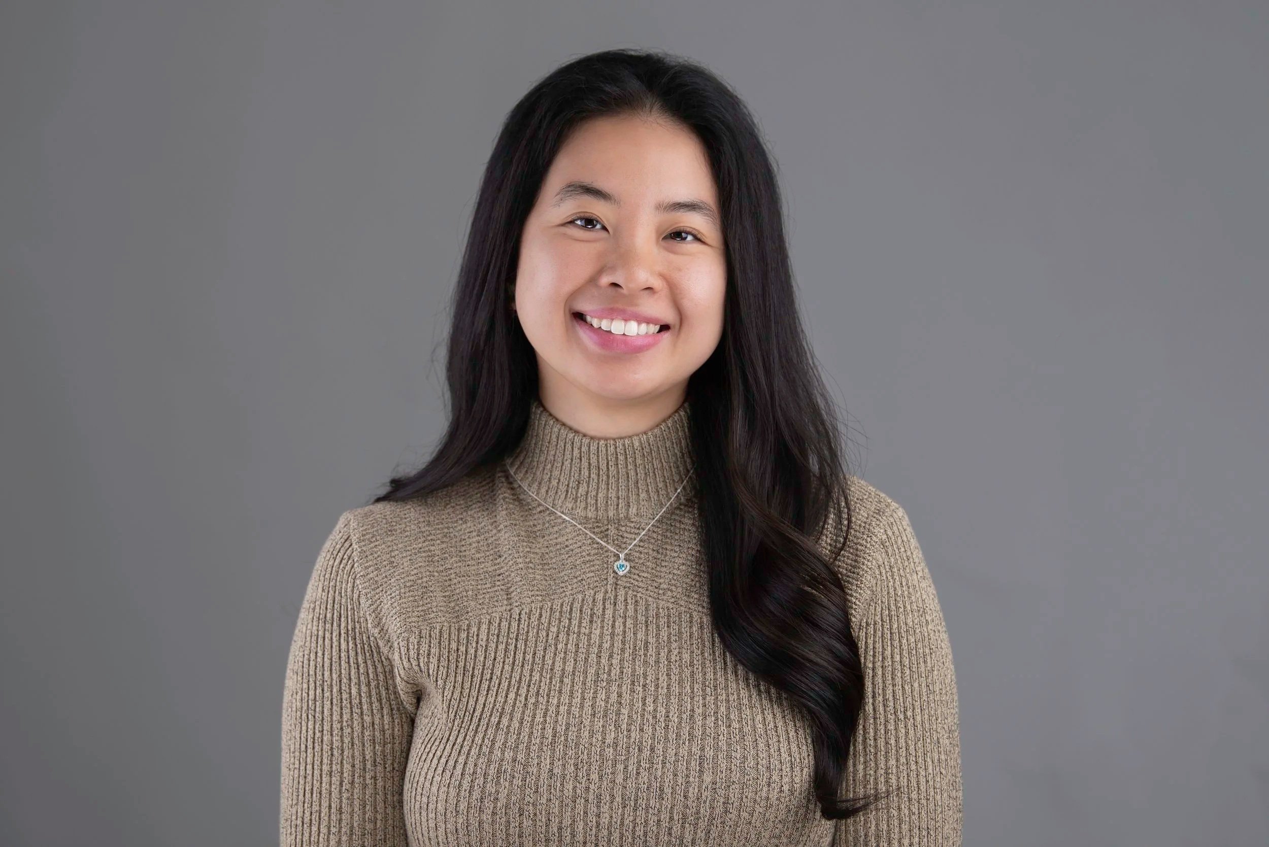A young Asian woman with long, dark hair smiling, wearing a beige ribbed sweater, a silver necklace with a heart-shaped pendant, against a plain gray background l l Encompass Growth Counselling l Lyn Le