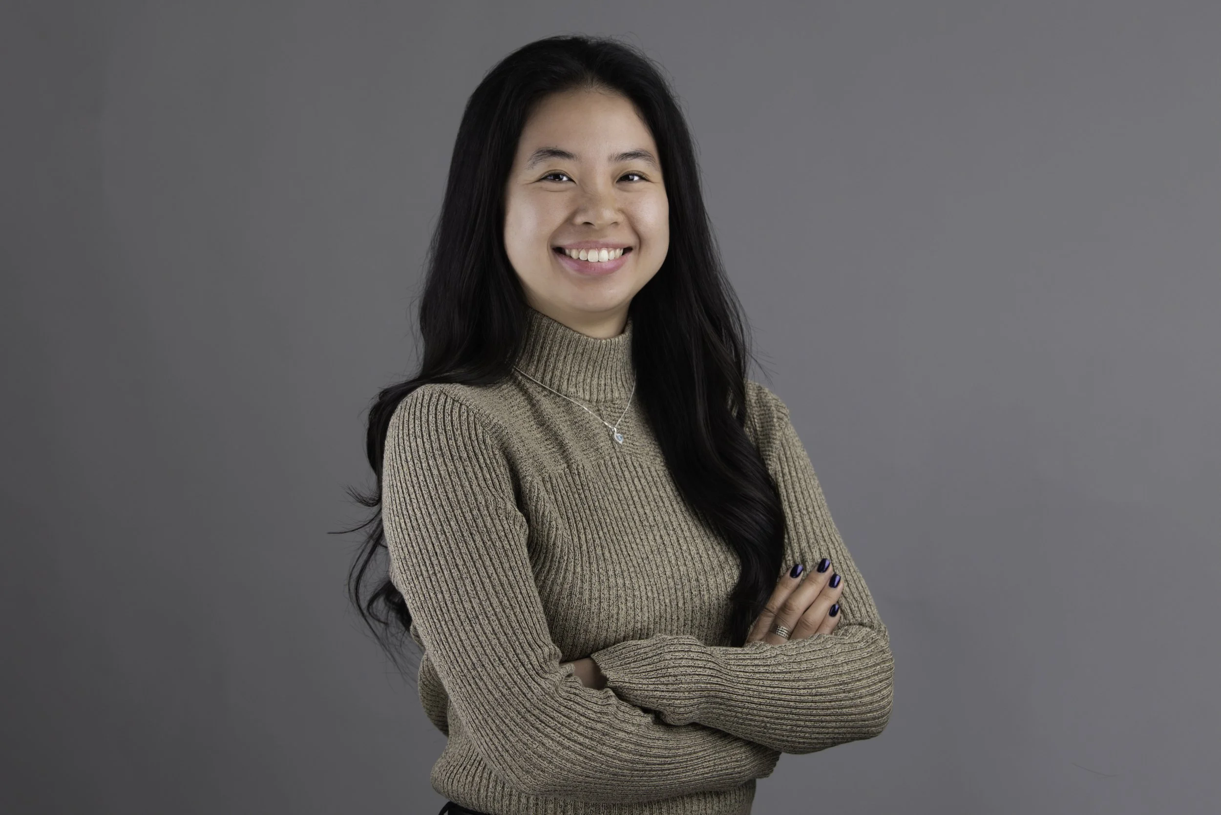 A young woman with long black hair, smiling, wearing a beige ribbed turtleneck sweater, silver necklace, and painted nails, standing against a plain gray background l Encompass Growth Counselling l Lyn Le