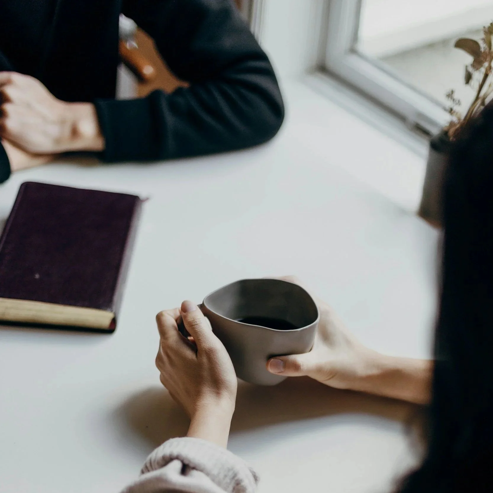 Person holding a coffee mug over a white table, with an open notebook and a person sitting across from them, wearing a black jacket. A glass door is visible in the background l Encompass Growth Counselling l Lyn Le