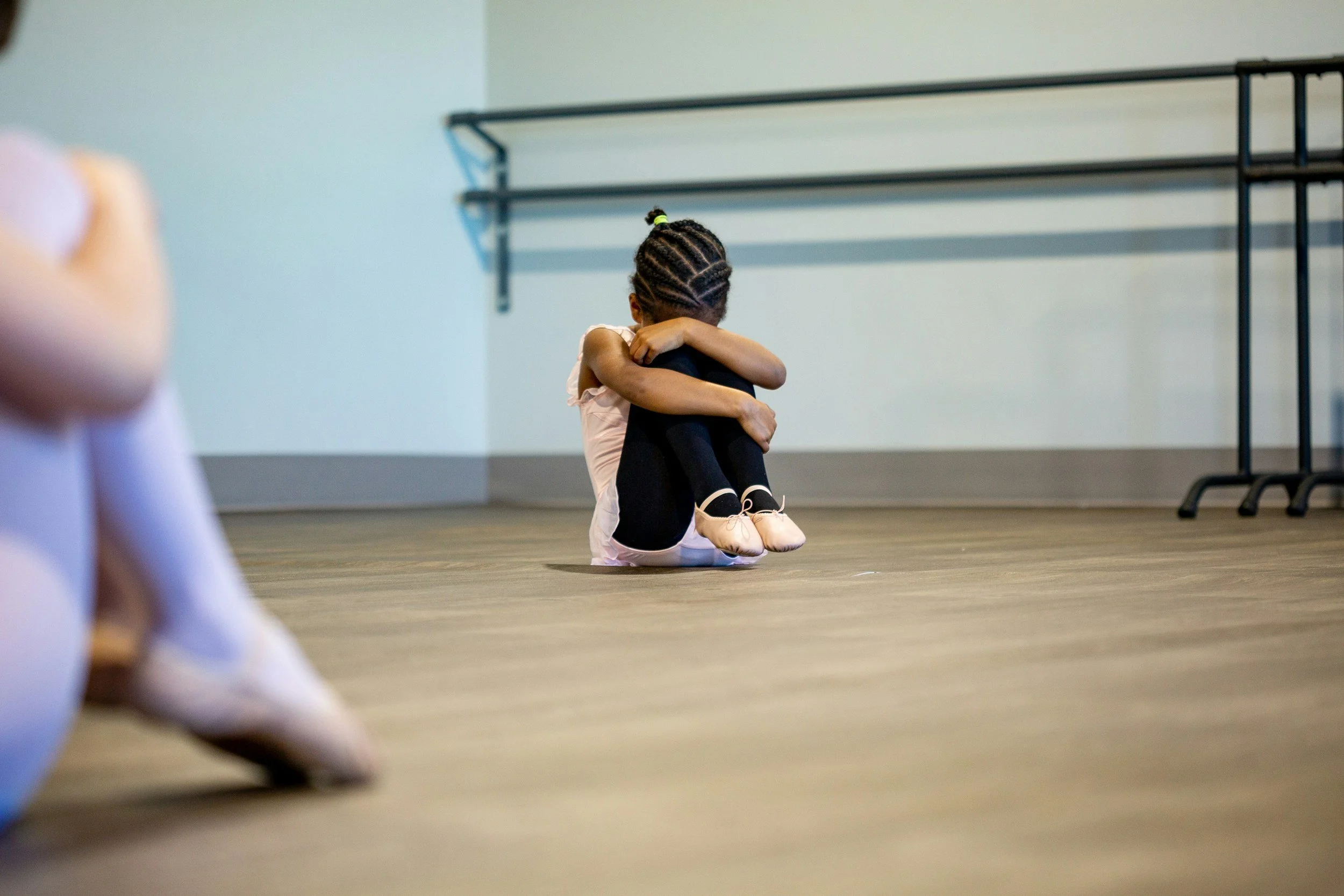 A young girl sitting on the floor with her knees pulled to her chest, hugging her legs, in a dance studio or rehearsal space.