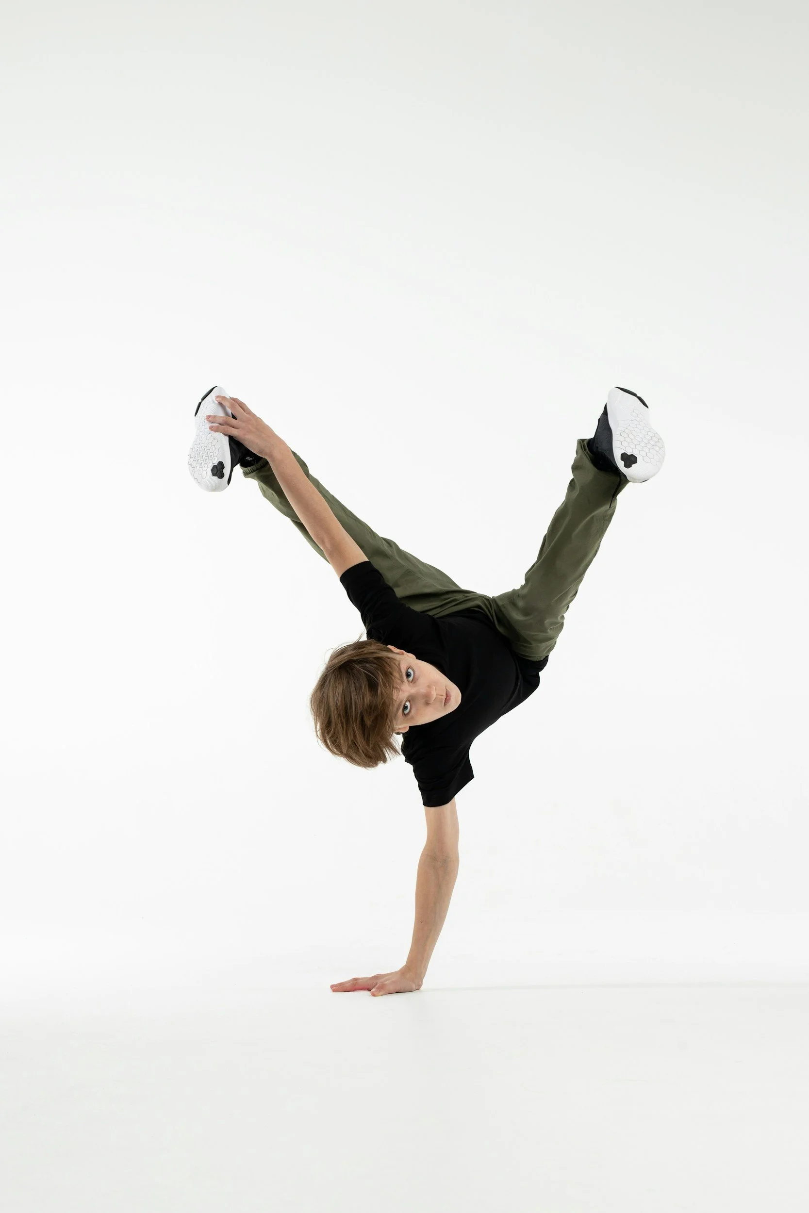 A young man performing a one-handed handstand with his legs spread wide apart, against a plain white background.