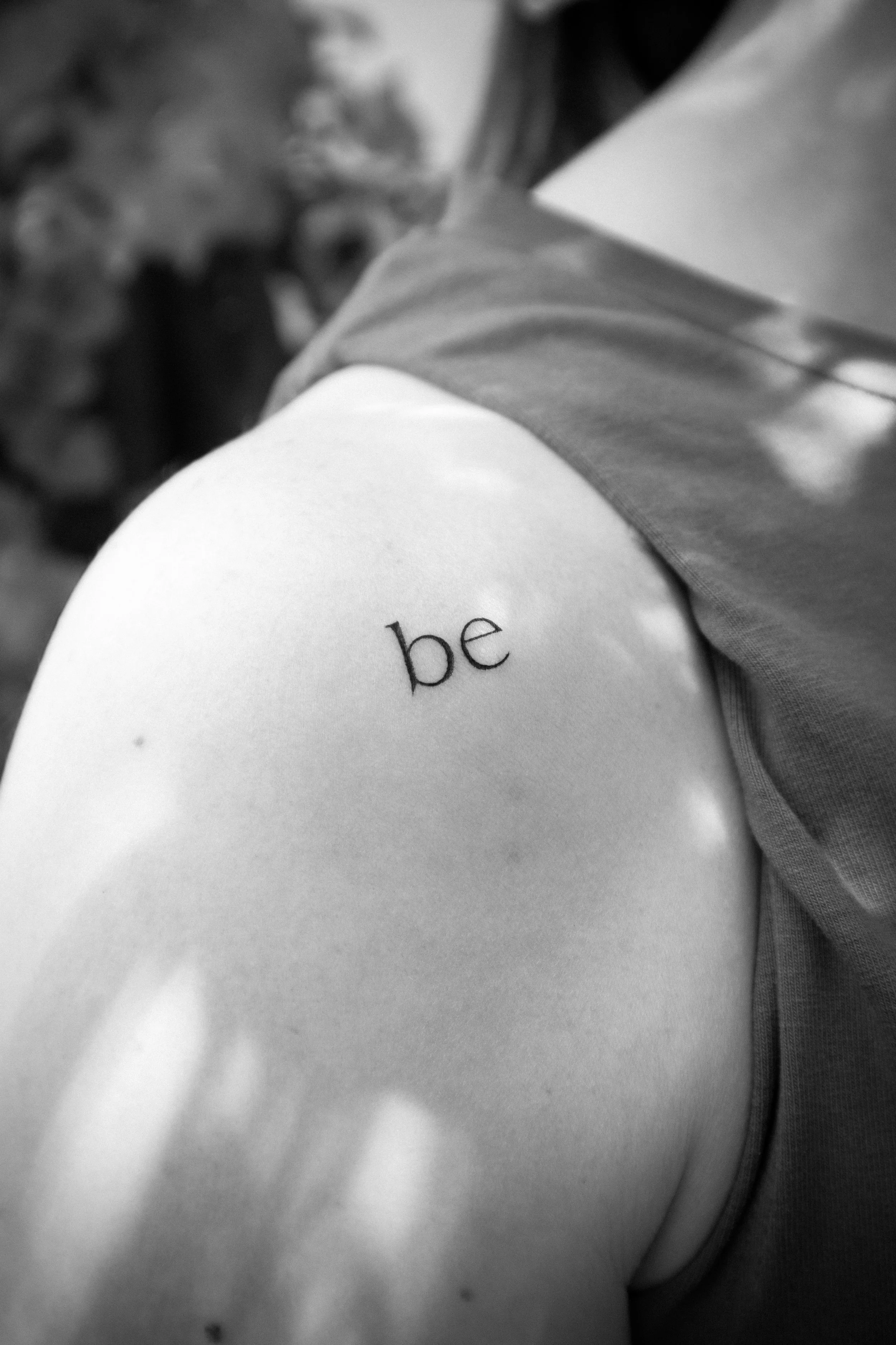 Tattoo en la parte superior del hombro que dice 'be' en letras pequeñas.